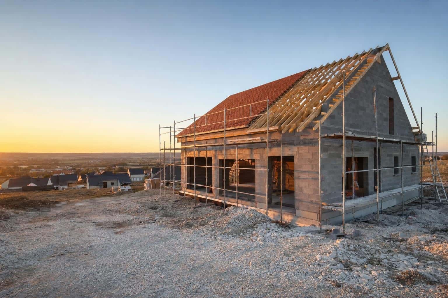 Maison neuve en construction aux murs en blocs béton, charpente bois et toiture en partie couverte de tuiles, au soleil couchant