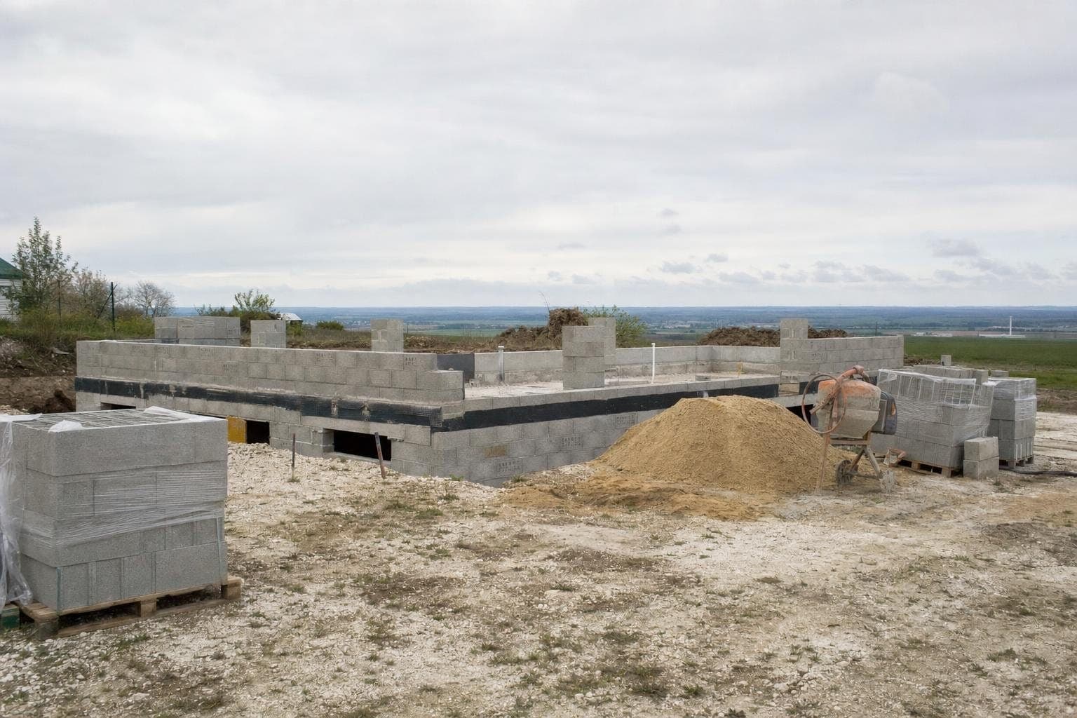 Chantier de maison avec soubassement en blocs béton, palettes de blocs et bétonnière sur un sol crayeux